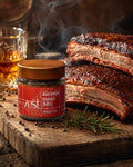 A ZEST Bourbon Barrel BBQ rub jar on a rustic wooden board, flanked by smoky barbecued ribs, a sprig of rosemary, scattered peppercorns, and a glass of whiskey.