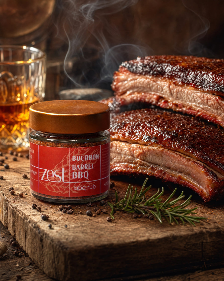 A ZEST Bourbon Barrel BBQ rub jar on a rustic wooden board, flanked by smoky barbecued ribs, a sprig of rosemary, scattered peppercorns, and a glass of whiskey.