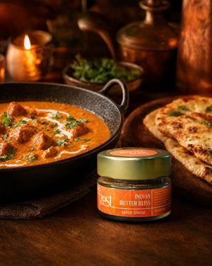 A ZEST Indian Butter Bliss spice blend jar displayed next to a rich butter chicken or paneer dish and naan bread on a dark wooden table, with candlelight in the background.